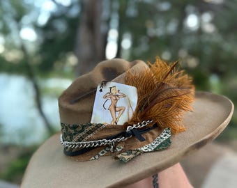 Puede incluir: Sombrero de vaquero color canela con una banda decorativa, una carta de juego y plumas marrones. La carta de juego presenta una ilustración pin-up. Una cadena plateada y tela estampada añaden detalles.