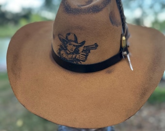 Puede incluir: Un sombrero de vaquero marrón con una banda de cuero negro. El sombrero presenta un gráfico de una mujer sosteniendo un arma y está adornado con una pluma y un elemento decorativo. El sombrero se exhibe al aire libre.