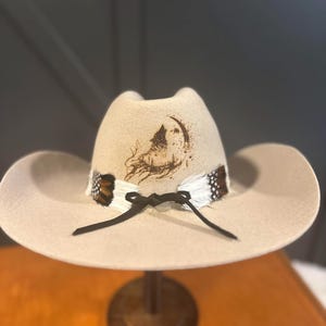 May include: A beige cowboy hat with a decorative band of feathers and a black tie. The hat features a detailed illustration of a wolf howling at the moon. The hat is displayed on a wooden stand.
