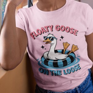 May include: Pale pink t-shirt featuring the text "FLOATY GOOSE ON THE LOOSE" and a cartoon goose wearing sunglasses and floating on a blue and black inflatable ring. The goose has yellow webbed feet. The shirt is being worn.