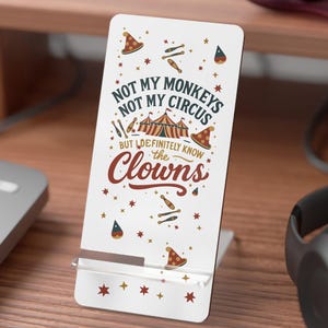 May include: A white phone stand with the text "Not My Monkeys Not My Circus But I Definitely Know the Clowns." The stand has a circus theme with illustrations of circus tents, hats, and stars. The stand is on a wooden desk.