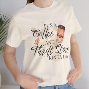 May include: Cream-colored t-shirt with the text "It's a Coffee and Thrift Store Kinda Day." The design includes a coffee cup illustration and a price tag. The shirt is short-sleeved and made of a soft material.