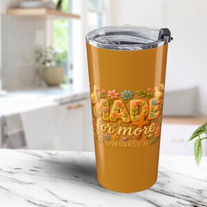 Op de afbeelding: Een goudgele reisbeker met een doorzichtig deksel. De beker heeft de tekst "MADE for MORE" in een bloemenontwerp, met de Bijbeltekst Efeziërs 2:10 eronder. De beker is ontworpen voor warme of koude dranken.
