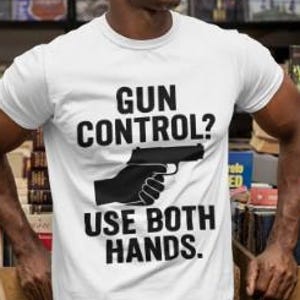 以下が含まれることがあります： 「GUN CONTROL？USE BOTH HANDS.」の黒い文字が入った白いTシャツ。グラフィックには黒い拳銃と銃を握る手が含まれています。画像内の人物はサングラスをかけています。