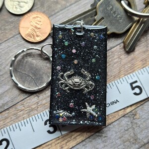 Op de afbeelding: Zwarte glitterhars sleutelhanger met een zilveren krabbe hanger en iriserende glitter accenten. De sleutelhanger is bevestigd aan een zilveren ring.
