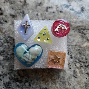 May include: Five small resin ornaments with different shapes and designs. A blue diamond with a silver cross, a yellow triangle with glitter, a red circle with a silver dolphin, a blue heart with seashells, and a pink square with a gold star.