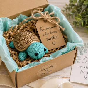 May include: A crocheted turtle with a gold shell and turquoise body inside a cardboard gift box. The box contains brown paper filler, light blue tissue paper, and a brown tag with the text For someone who loves turtles and a heart symbol.