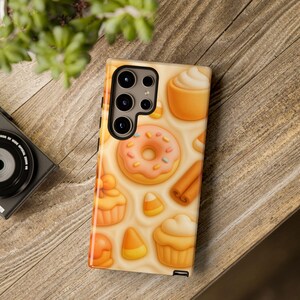 Puede incluir: Una funda de teléfono con un patrón repetido de productos horneados. El diseño incluye una rosquilla rosa glaseada con chispas, magdalenas, rollos de canela y caramelos de maíz sobre un fondo color crema.