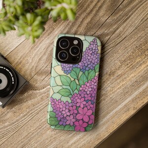 May include: A smartphone case with a floral pattern of purple and pink flowers, green leaves, and a light green backdrop. The case is on a wooden surface with a camera and plant in the background.