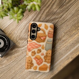 Puede incluir: Una funda de teléfono con un diseño temático de picnic. La funda presenta ilustraciones de una cesta de picnic, osos, bellotas, hojas, un pastel y una manta a cuadros en tonos marrones, naranjas y beige. La funda está sobre una superficie de madera.