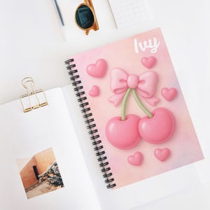May include: A spiral-bound notebook with a pink cover featuring a 3D illustration of pink cherries, a bow, and hearts. The word "Ivy" is written in cursive. The notebook is open on a white surface.