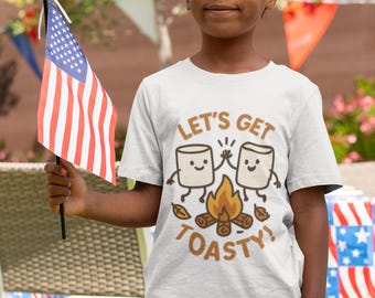 Op de afbeelding: Een kind draagt een lichtgrijs t-shirt met de tekst "LET'S GET TOASTY!" en een afbeelding van twee marshmallows die boven een kampvuur roosteren. De persoon houdt een kleine Amerikaanse vlag vast.