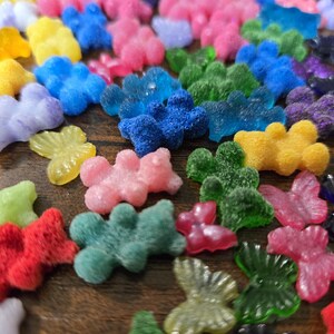 May include: A collection of small, colourful bear and butterfly shaped charms in various textures, including translucent, glittery, and fuzzy finishes. The assortment features vibrant shades of red, blue, green, yellow, pink, and purple scattered across the surface.