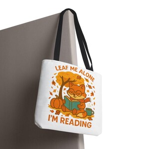 Puede incluir: Bolso tote blanco con asas negras. Presenta un zorro de dibujos animados leyendo un libro bajo un árbol con hojas cayendo. El texto "LEAF ME ALONE I'M READING" está impreso en el bolso, junto con una calabaza y una taza de café.