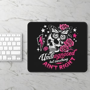Op de afbeelding: Zwarte muismat met een schedel-, vlinder- en rozenontwerp. De tekst luidt "Undiagnosed but something ain't right" in roze en wit. Het ontwerp bevat sterren en bliksemschichten.