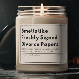 May include: A beige candle in a clear glass jar with a black label that reads "Smells like Freshly Signed Divorce Papers" and a smaller text that reads "Minded with a hint of freedom and the knowledge it's not your fucking problem anymore".
