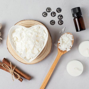 May include: A flat lay of candle making supplies including white soy wax flakes in a heart-shaped bowl, a wooden spoon with wax flakes, two white candles, cinnamon sticks, twine, and a small bottle of essential oil.