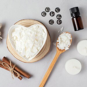 May include: A flat lay of candle making supplies including white wax flakes in a heart-shaped bowl, a wooden spoon with wax flakes, two white candles, a spool of twine, cinnamon sticks, and a small bottle of essential oil.