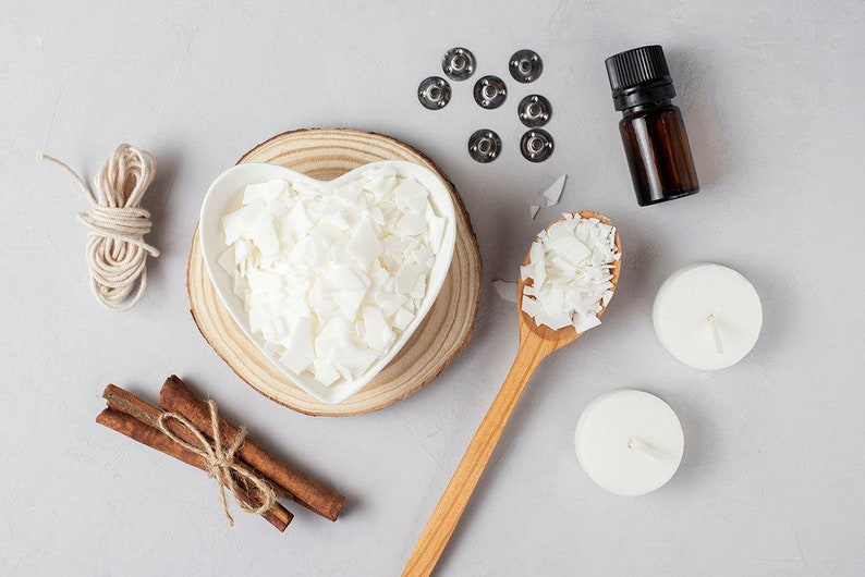 May include: A flat lay of candle making supplies including white wax flakes in a heart-shaped bowl, a wooden spoon with wax flakes, two white candles, a spool of twine, cinnamon sticks, and a small bottle of essential oil.