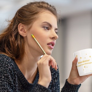 Puede incluir: Una mujer sostiene una taza blanca con letras doradas que dicen "Rockstar Program Manager" y estrellas doradas. Está pensando mientras se sostiene un lápiz en la mejilla.