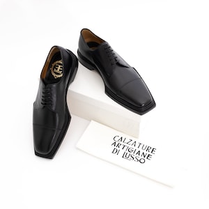 May include: Two black leather lace-up shoes with square toes. The shoes are on a white surface with a white box and a white card that says "Calzature Artigiane Di Lusso".