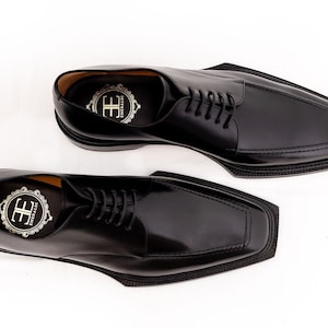 May include: A pair of black leather dress shoes with a unique square toe design. The shoes have a lace-up closure and a glossy finish. The insoles feature a gold emblem. The shoes are set against a white background.