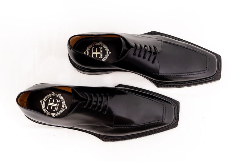 May include: A pair of black leather dress shoes with a unique square toe design. The shoes have a lace-up closure and a glossy finish. The insoles feature a gold emblem. The shoes are set against a white background.