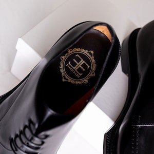 May include: Close-up of a pair of black leather dress shoes. The interior of one shoe reveals a gold emblem with the word "EMERALDI" and the initials "EE". The shoes have a glossy finish and are displayed on a white surface.