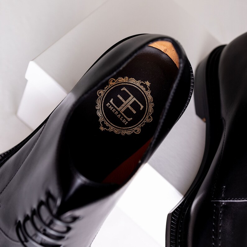 May include: Close-up of a pair of black leather dress shoes. The interior of one shoe reveals a gold emblem with the word "EMERALDI" and the initials "EE". The shoes have a glossy finish and are displayed on a white surface.
