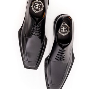 May include: A pair of black leather dress shoes with square toes and lace-up closures. The shoes have the brand name "EMERALDI" printed on the insole. The shoes are set against a white background.