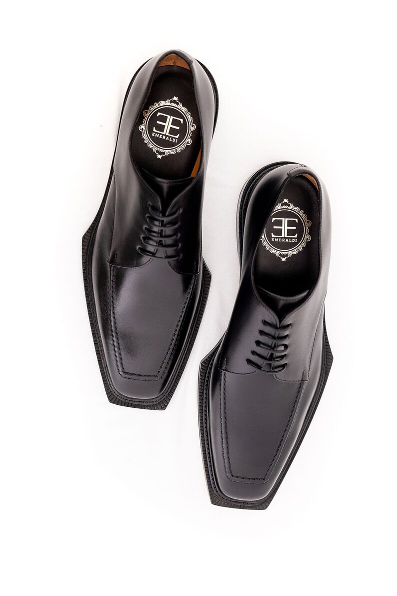 May include: A pair of black leather dress shoes with square toes and lace-up closures. The shoes have the brand name "EMERALDI" printed on the insole. The shoes are set against a white background.