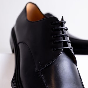 May include: Close-up of a pair of black leather dress shoes. The shoes have a classic design with a rounded toe, a lace-up closure, and a thick sole. The leather appears smooth and polished, with visible stitching details.