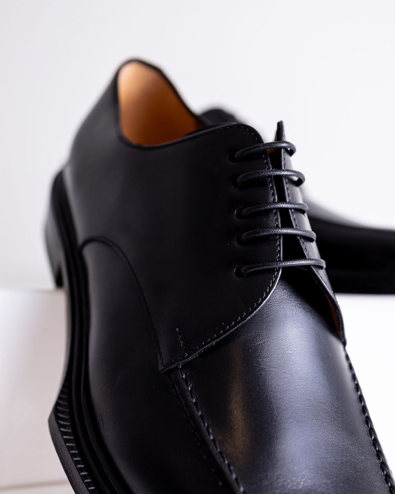 May include: Close-up of a pair of black leather dress shoes. The shoes have a classic design with a rounded toe, a lace-up closure, and a thick sole. The leather appears smooth and polished, with visible stitching details.