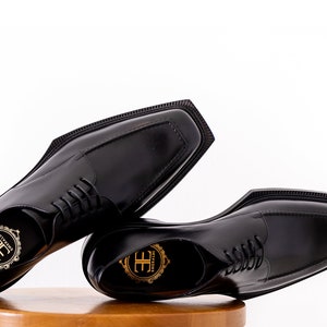 May include: A pair of black leather dress shoes with a square toe design. The shoes have a lace-up closure and a small logo on the insole. The shoes are displayed on a wooden surface against a white background.