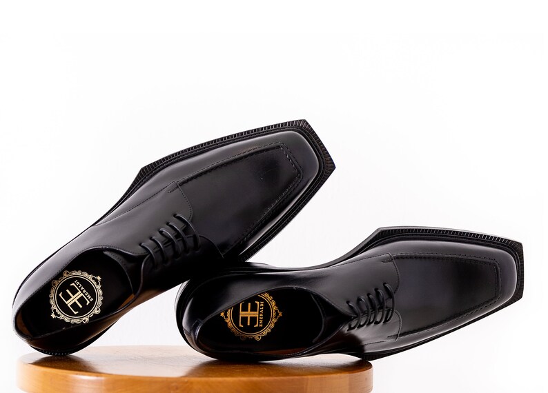 May include: A pair of black leather dress shoes with a square toe design. The shoes have a lace-up closure and a small logo on the insole. The shoes are displayed on a wooden surface against a white background.