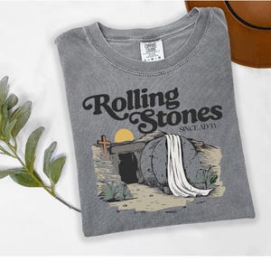 May include: Grey t-shirt featuring the text "Rolling Stones Since AD 33". The graphic shows a tomb with a cross and a sun rising. The t-shirt is folded on a white surface.