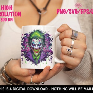 May include: A white mug featuring a colourful illustration of the Joker from Batman. The Joker is depicted with green hair, purple and green clothing, and a dripping green smile. The background is a purple and green swirl.