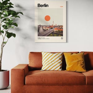 Puede incluir: Un p&oacute;ster enmarcado que presenta una imagen de estilo vintage de Berl&iacute;n, Alemania, con un gran sol naranja. La impresi&oacute;n incluye el nombre de la ciudad y las coordenadas. Se muestra sobre un sof&aacute; color &oacute;xido con cojines amarillos y blancos.