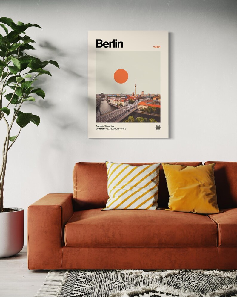 Puede incluir: Un p&oacute;ster enmarcado que presenta una imagen de estilo vintage de Berl&iacute;n, Alemania, con un gran sol naranja. La impresi&oacute;n incluye el nombre de la ciudad y las coordenadas. Se muestra sobre un sof&aacute; color &oacute;xido con cojines amarillos y blancos.