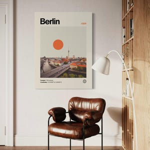 Puede incluir: Un p&oacute;ster enmarcado con la palabra "Berlin" en negro, un paisaje urbano y un gran c&iacute;rculo naranja. El p&oacute;ster est&aacute; sobre un sill&oacute;n de cuero marr&oacute;n con patas de metal negro. El texto "/GER" tambi&eacute;n est&aacute; incluido.