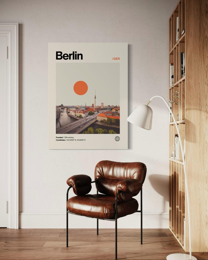 Puede incluir: Un p&oacute;ster enmarcado con la palabra "Berlin" en negro, un paisaje urbano y un gran c&iacute;rculo naranja. El p&oacute;ster est&aacute; sobre un sill&oacute;n de cuero marr&oacute;n con patas de metal negro. El texto "/GER" tambi&eacute;n est&aacute; incluido.