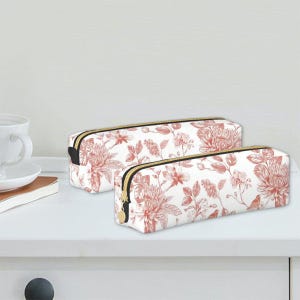 Puede incluir: Dos estuches blancos con un estampado floral rojo. Los estuches tienen cremalleras negras con tiradores dorados. Los estuches son rectangulares y están sobre una superficie blanca.