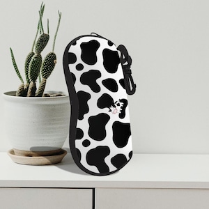 Puede incluir: Estuche para gafas con estampado de vaca en blanco y negro, con una correa negra y un pequeño dibujo animado de una vaca. El estuche está diseñado para proteger las gafas y está hecho de un material duradero.