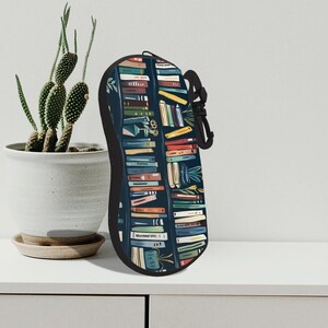 Può includere: Un astuccio nero per occhiali con un colorato design a libreria. L'astuccio è dotato di chiusura a zip e di un anello nero per il trasporto. Il design include vari titoli di libri ed elementi decorativi su sfondo blu scuro.