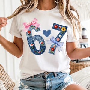 Pode incluir: Camiseta branca com os números "6" e "7" aplicados em denim, decorados com bordados florais e laços. Um pequeno coração azul está entre os números. A camiseta é combinada com shorts jeans.