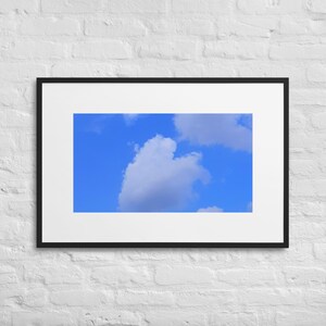 May include: Framed art print featuring a bright blue sky with fluffy white and gray clouds. The artwork is presented in a black frame with a white mat, ready to hang. The print is mounted on a white brick wall.