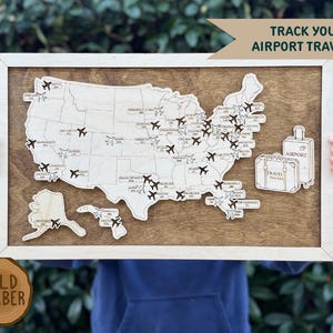 May include: Wooden map of the United States with airplane cutouts marking airport locations. The map is set in a wooden frame with a dark brown background. The text "TRACK YOUR AIRPORT TRAVELS!" is displayed above the map. Two suitcase illustrations are on the right.