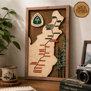 May include: A rectangular wooden wall art piece featuring a light-coloured wood map of the Appalachian Trail inside a dark wood frame. The map includes a green Appalachian Trail logo, state abbreviations, a black trail line, and red and green markers.