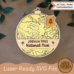 May include: Wooden ornament with a topographical map of Joshua Tree National Park. The ornament has a yellow background with brown contour lines and the park's name. A wooden emblem with a tree is on the top right. The text "Actual Park Topography!" is also visible.