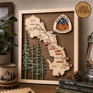 May include: A framed wooden wall art piece featuring a laser-cut map of the John Muir Trail. The map displays various landmarks, national parks, and wilderness areas in light wood against a dark wood background, with green pine tree cutouts.